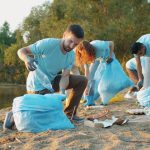 Grupo de voluntários recolhe lixo às margens de um rio usando luvas e sacos para preservar o ambiente.