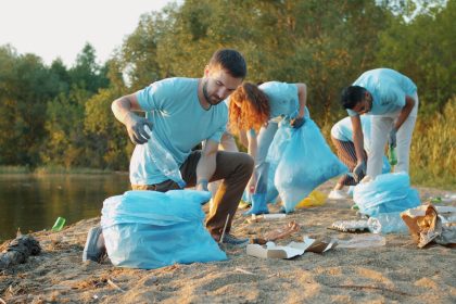 Grupo de voluntários recolhe lixo às margens de um rio usando luvas e sacos para preservar o ambiente.