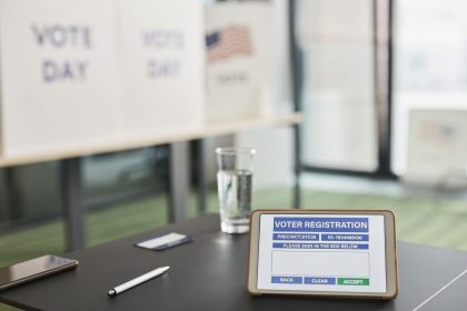 Tablet exibindo um formulário de registro eleitoral em uma estação de votação interna, com sinalização sobre o dia da eleição.